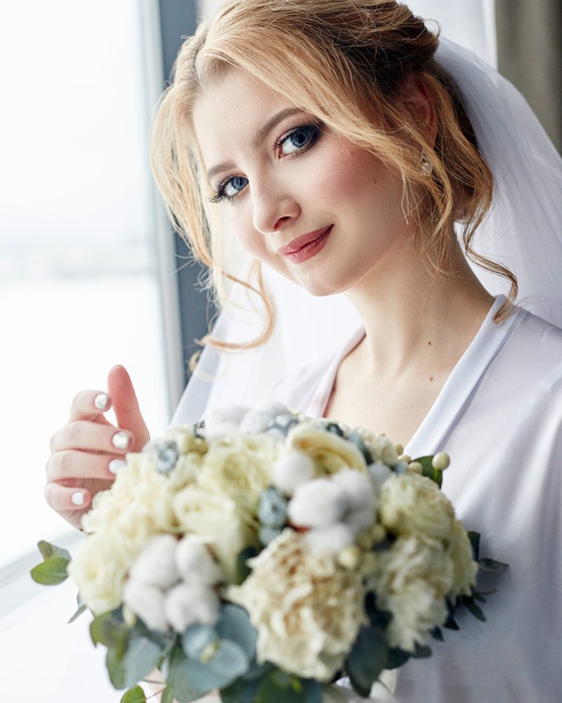 Morning portrait of the bride preparing for the wedding ceremony. Portrait of a blonde woman near the window in a white robe and a veil on her head